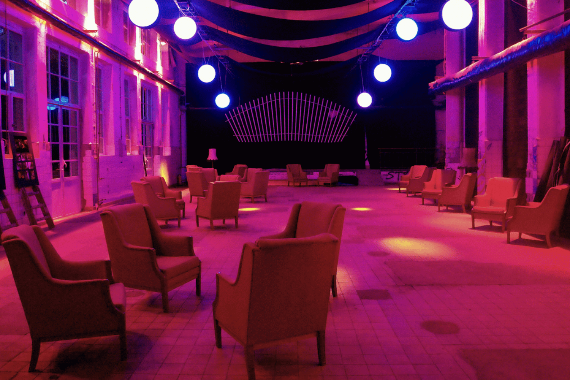 Ein stimmungsvoll beleuchteter, großer Veranstaltungsraum in einem industriellen Gebäude. Der Raum ist in intensives pinkes und violettes Licht getaucht. Im gesamten Bereich sind zahlreiche beige Polstersessel locker verteilt, die zum Verweilen einladen. Von der hohen Decke hängen mehrere große, hell leuchtende Kugel-Lampions sowie schwarze Stoffbahnen, die bogenförmig gespannt sind. Im Hintergrund befindet sich eine schwarze Bühne mit einer fächerartigen, weißen Holzkonstruktion als Kulisse. Die großen Fenster auf der linken Seite lassen das violette Licht nach außen dringen und unterstreichen die besondere Atmosphäre des Ortes.