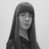 Ein Schwarz-Weiß-Porträt einer jungen Frau mit langem, glattem dunklem Haar und Pony. Sie trägt eine Brille und ein dunkles Oberteil und blickt mit neutralem Gesichtsausdruck in die Kamera.