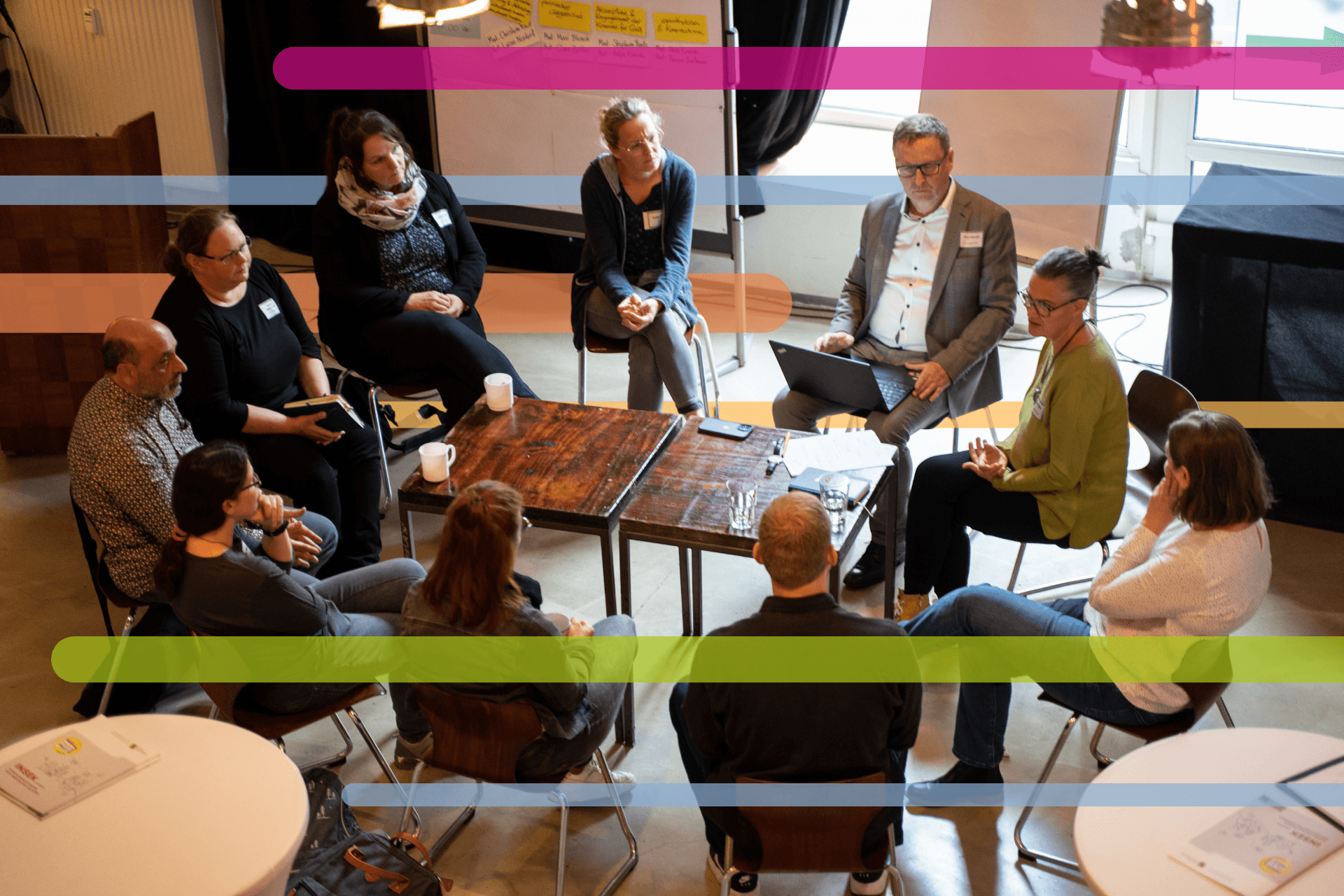 Eine Gruppe von Menschen sitzt bei einem Workshop im Kreis um mehrere Tische und diskutiert miteinander; einige machen Notizen oder arbeiten am Laptop. Im Hintergrund sind Pinnwände und Moderationsmaterialien zu sehen.
